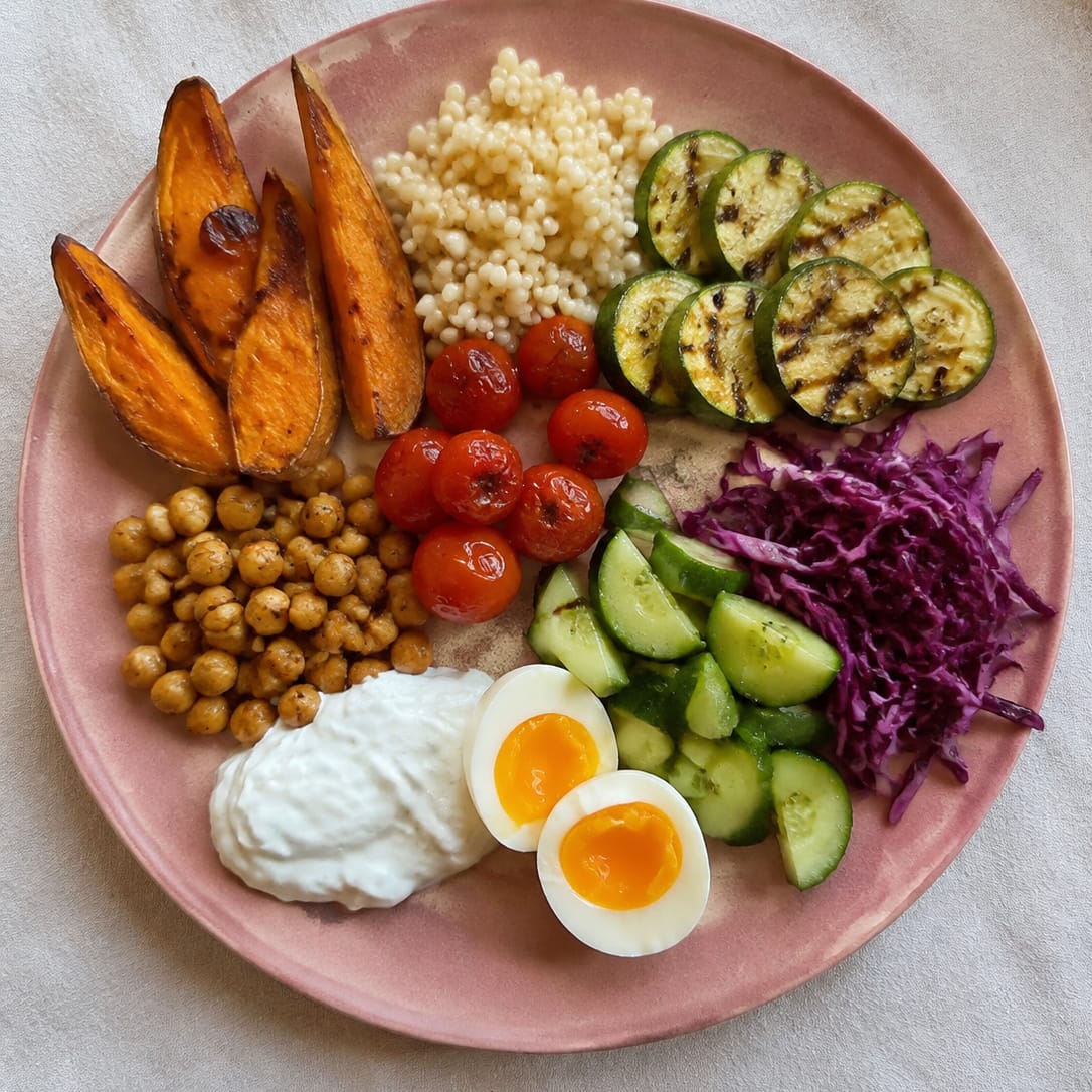 A plate seen from above: roast sweet potato wedges, pearl couscous, spiced roast chickpeas, grilled courgette rounds, blistered cherry tomatoes, chopped cucumber, shredded red cabbage, a spoonful of yoghurt and a halved soft-boiled egg, on a pink ceramic plate on linen.
